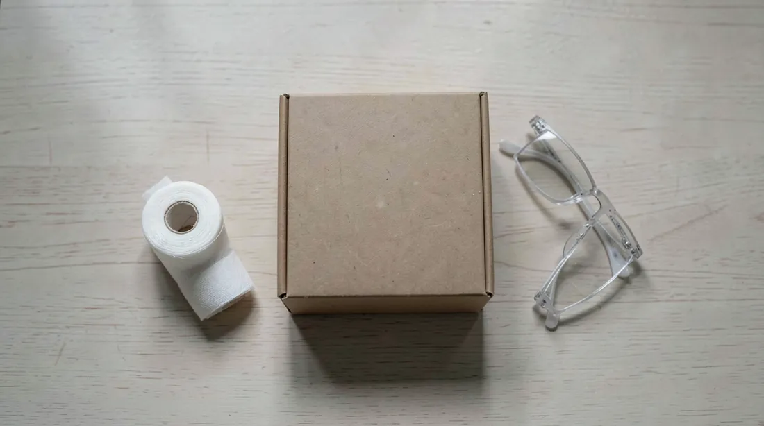 Top-down view of a bare light wooden table with a single plain closed brown cardboard box with blank sides in the centre, a roll of white gauze bandage and a pair of folded reading glasses beside it